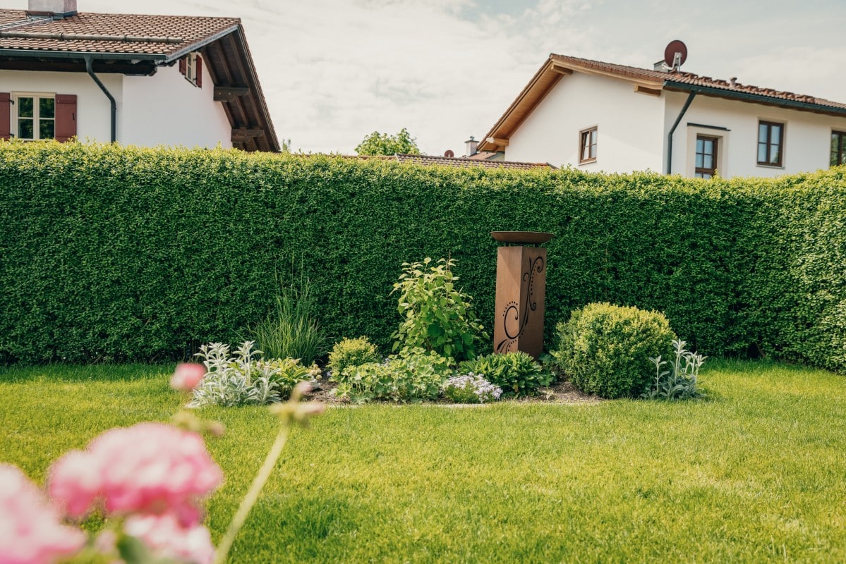 Eine ordentlich gestutzte grüne Hecke trennt zwei Häuser voneinander. Davor umgibt ein kleines Gartenbeet mit verschiedenen Pflanzen und Blumen einen dekorativen Holzpfosten. Rosafarbene Blumen sind im Vordergrund verschwommen zu erkennen.
