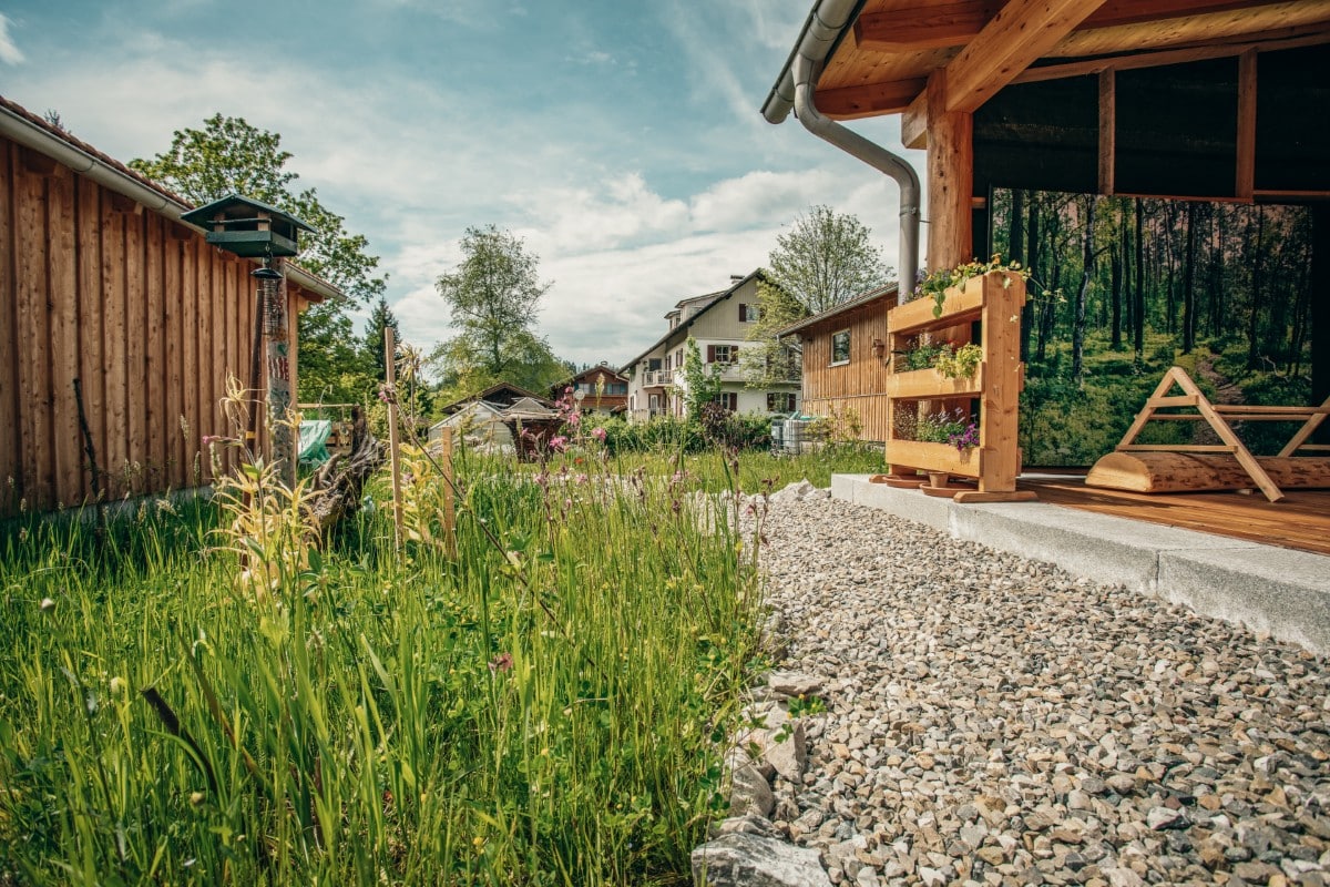 Eine kleine Hütte mit einer hölzernen Veranda und einem Kiesweg blickt auf einen Garten mit hohem Gras und Blumen; im Hintergrund sind Häuser und Bäume unter einem teilweise bewölkten Himmel zu sehen.