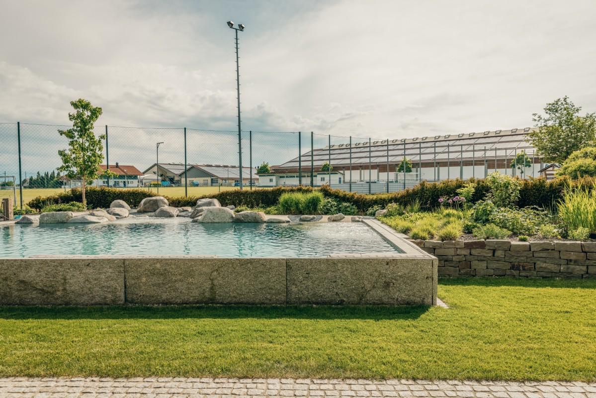 Ein rechteckiger Swimmingpool im Freien mit Felsen auf einer Seite, umgeben von Gras und Landschaftspflanzen, in der Nähe eines Sportplatzes mit einem Zaun und Gebäuden im Hintergrund.