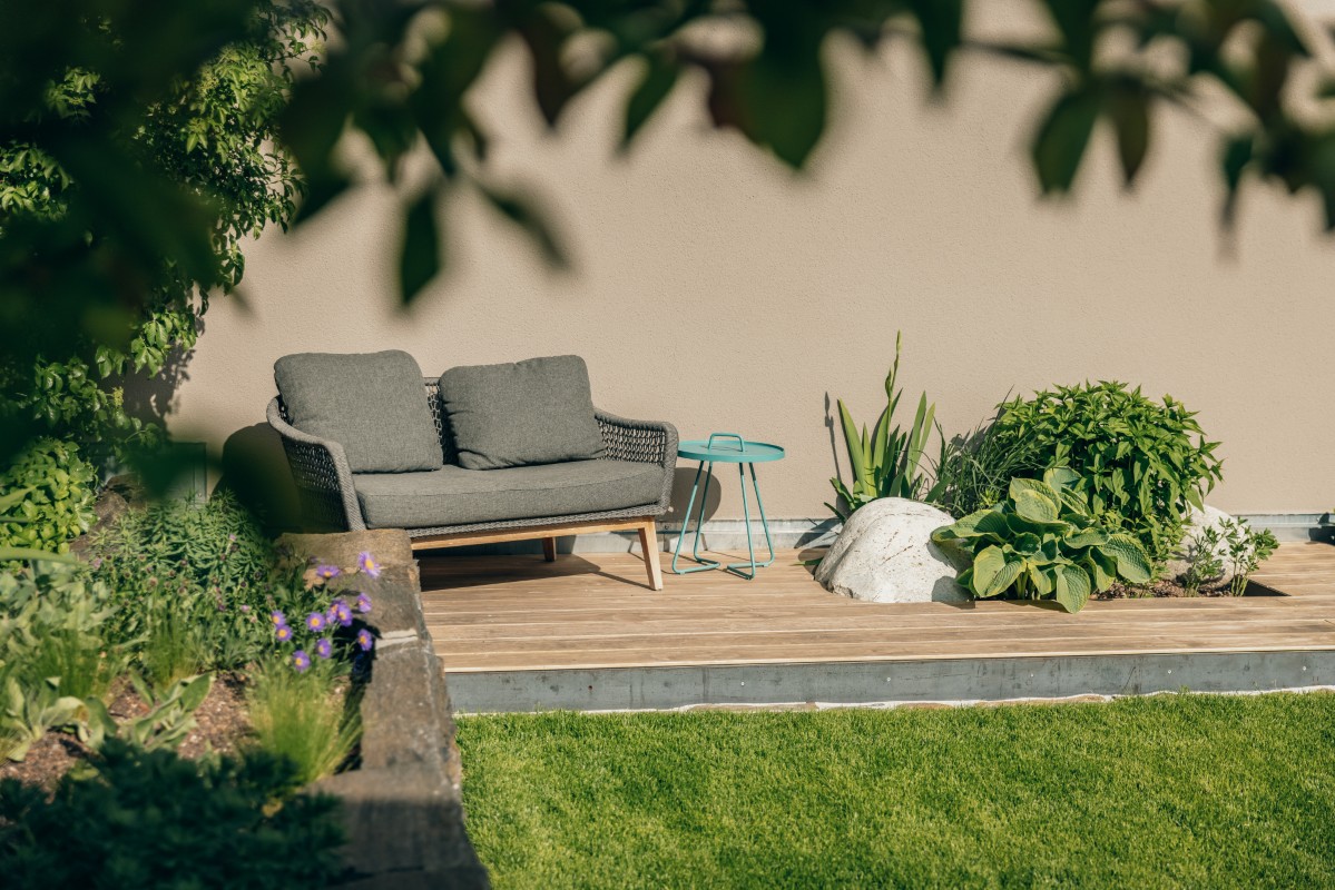 Auf der gemütlichen Terrasse stehen ein grauer, gepolsterter Liegesessel, ein kleiner blauer Beistelltisch und üppige Grünpflanzen auf einer Holzterrasse, die von einem Rasen umgeben ist und von überhängenden Ästen beschattet wird.