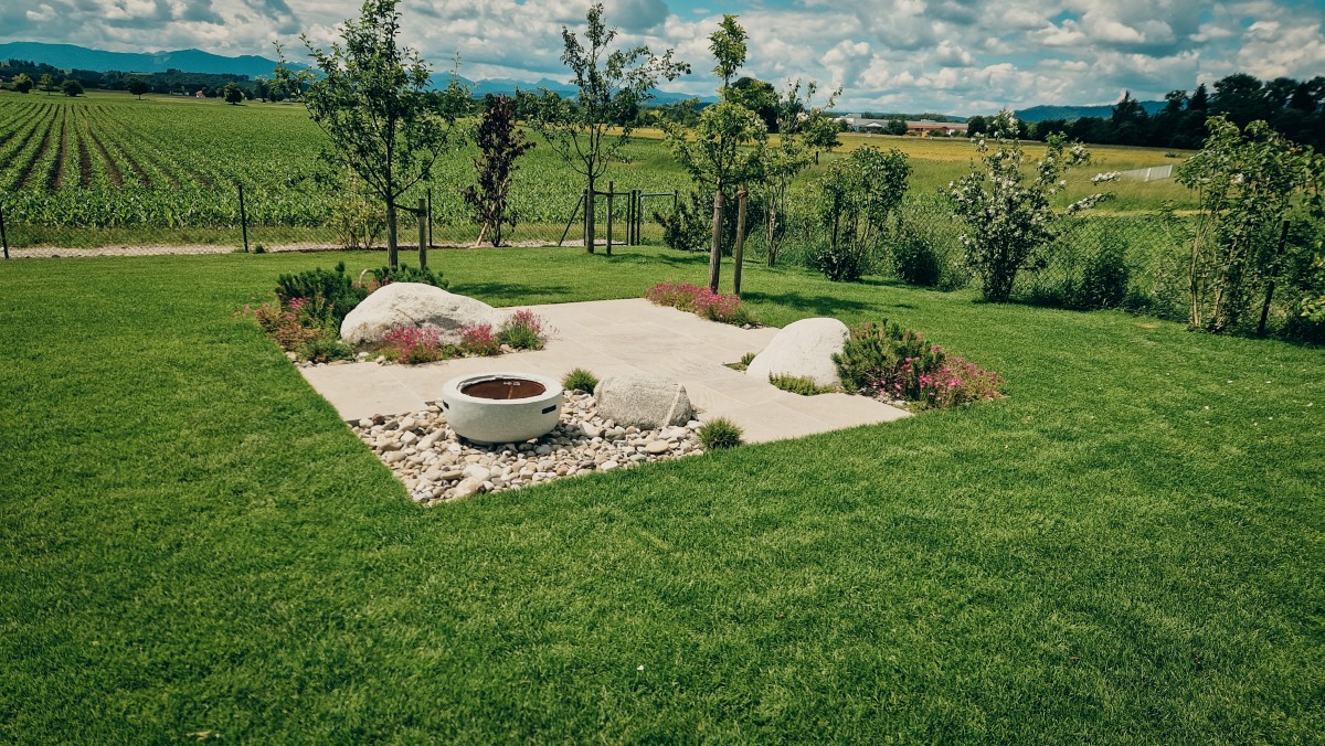 Eine Gartenlandschaft mit einer runden Feuerstelle, umgeben von Steinen, Kieselsteinen, Blumen und kleinen Bäumen, liegt in einem grasbewachsenen Hof mit Blick auf bewirtschaftete Felder und ferne Hügel unter einem teilweise bewölkten Himmel.