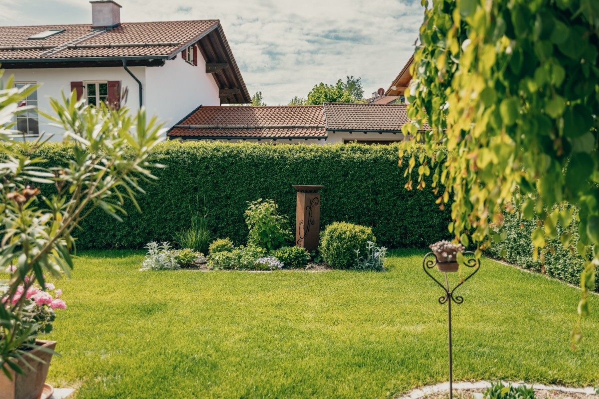 Ein gepflegter Hinterhofgarten mit grünem Gras, kleinen Pflanzen und einem dekorativen Holzpfosten in der Mitte. Hecken säumen die Rückseite, und hinter dem Garten ist ein weißes Haus mit braunen Zierleisten zu sehen.