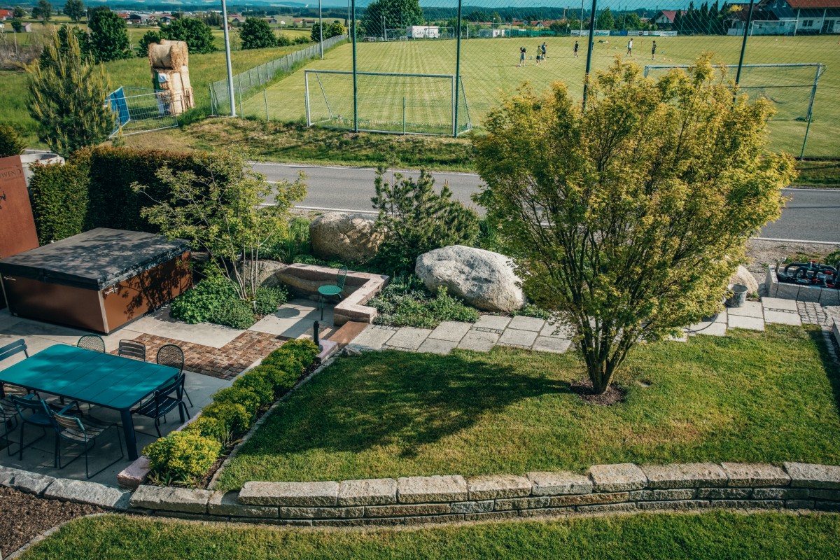 Ein begrünter Hinterhof mit Terrassenmöbeln, Bäumen und Sträuchern, der von einer Steinmauer begrenzt wird. Eine Straße trennt den Garten von einem Fußballplatz, auf dem Menschen spielen, während im Hintergrund Häuser und Hügel zu sehen sind.
