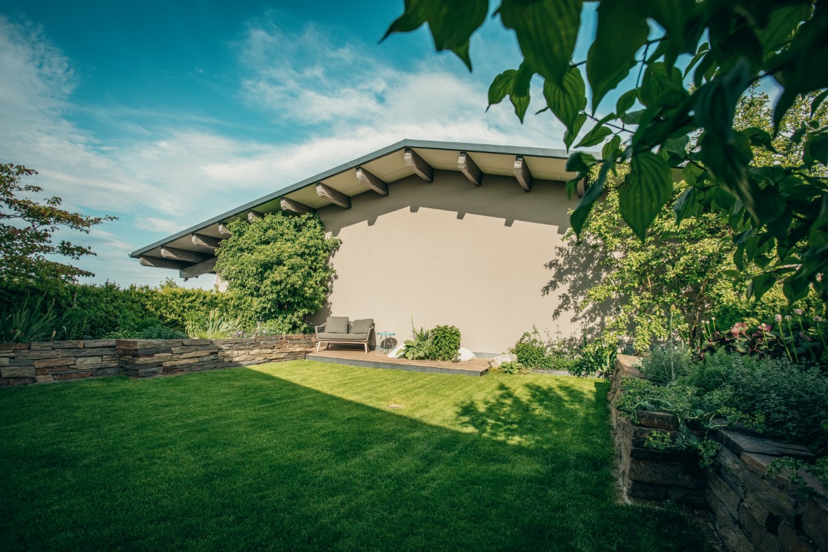 Ein üppig grüner Garten mit gepflegtem Rasen, mit Steinen eingefassten Gartenbeeten, Bäumen, Sträuchern und einem modernen Haus mit einer schattigen Terrasse und Sitzgelegenheiten im Freien unter einem blauen Himmel mit vereinzelten Wolken.