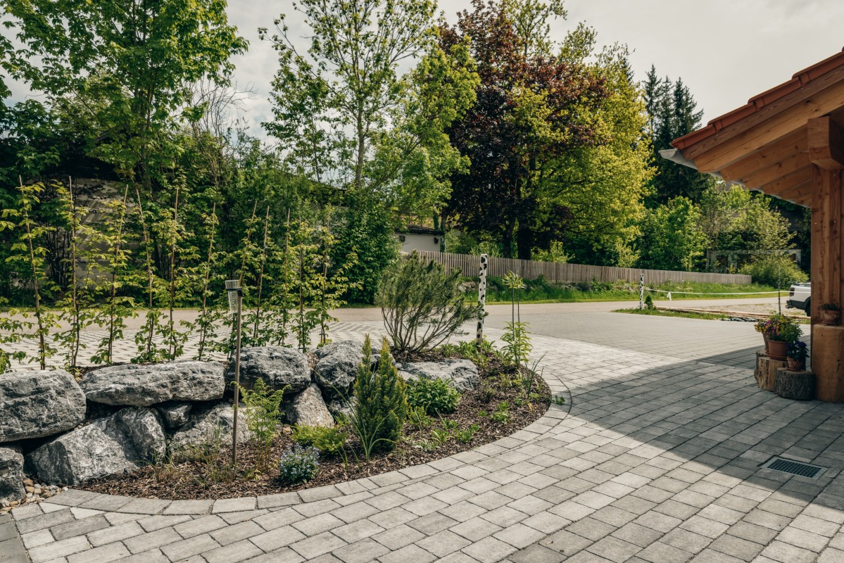 Eine gepflasterte Einfahrt schlängelt sich neben einem Landschaftsgarten mit Steinen und kleinen Pflanzen. Im Hintergrund sind Bäume und ein Holzzaun zu sehen, und rechts ist ein Teil eines Holzgebäudes zu erkennen. Die Szene ist hell und grün.