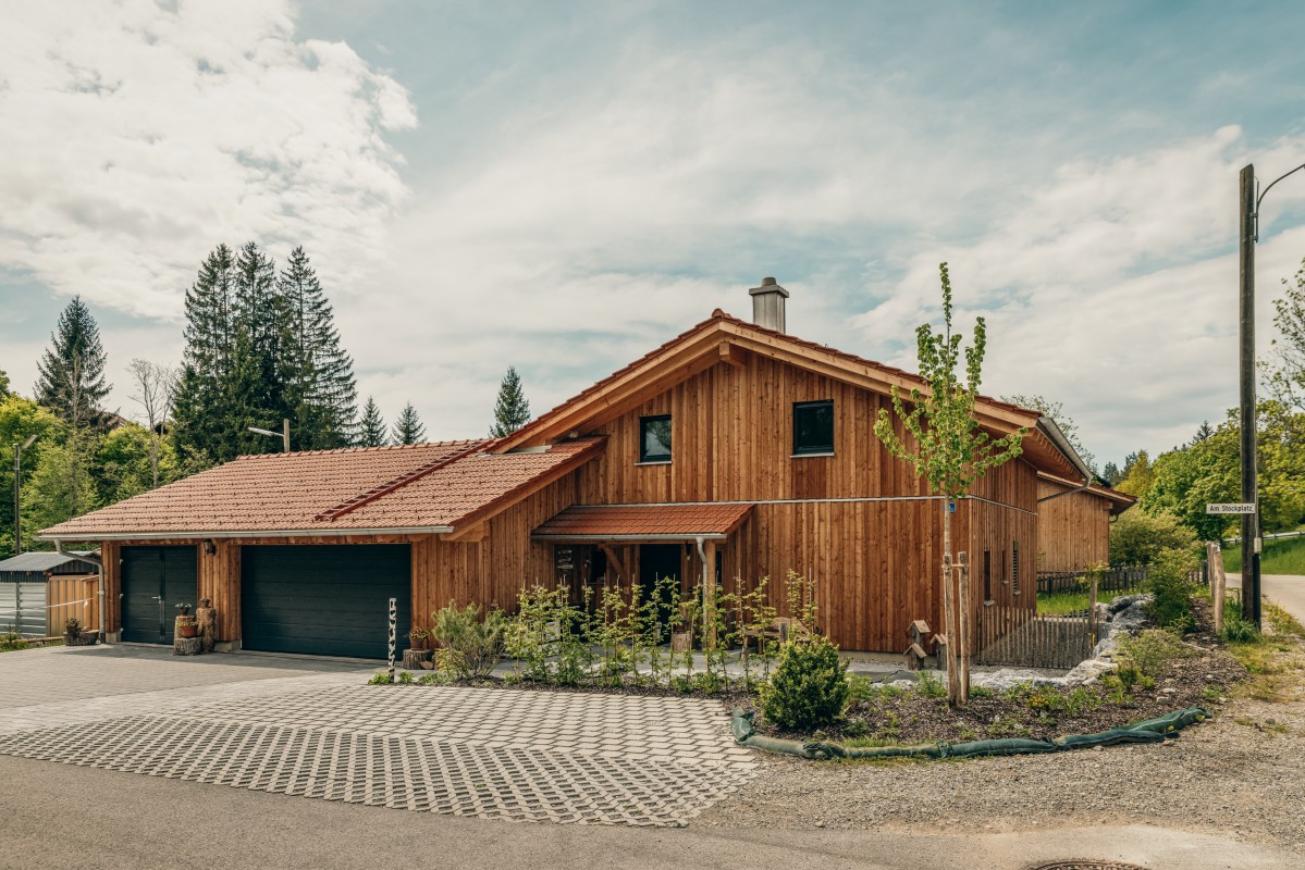 Ein modernes Holzhaus mit rotem Ziegeldach und schwarzen Garagentoren steht auf einem Eckgrundstück, umgeben von jungen Bäumen und Grünpflanzen, vor einem Hintergrund aus hohen Kiefern und einem teilweise bewölkten Himmel.