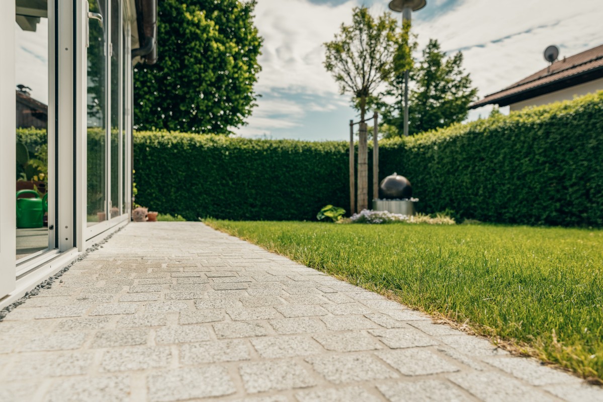 Ein sauber gepflasterter Steinweg führt entlang eines grünen Rasens in einem Hinterhof, der von gestutzten Hecken und kleinen Bäumen gesäumt wird, mit einem Haus und einem großen Fenster auf der linken Seite.