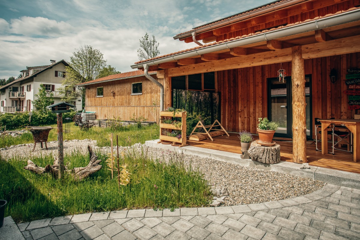 Ein rustikales Holzhaus mit einer überdachten Veranda, Topfpflanzen und Terrassenmöbeln, umgeben von einem grasbewachsenen Hof mit Gartendekorationen und einem gepflasterten Steinweg. Im Hintergrund sind andere Häuser und Bäume zu sehen.