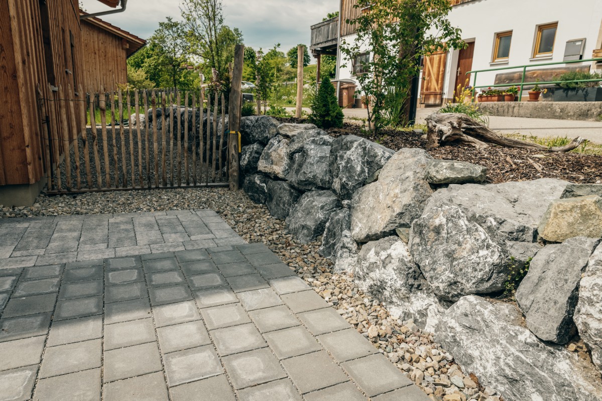 Eine gepflasterte Steinterrasse führt zu einem Holztor und einer Steinmauer. An einem sonnigen Tag sieht man im Hintergrund Häuser, Pflanzen und Bäume.