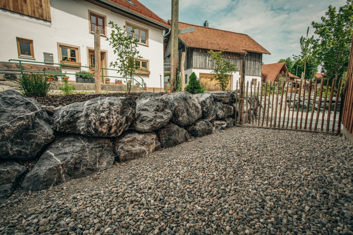 Ein Kiesweg führt an einer rustikalen Steinmauer und einem Holzzaun entlang, mit einem traditionellen Haus und einer Scheune im Hintergrund unter einem teilweise bewölkten Himmel.