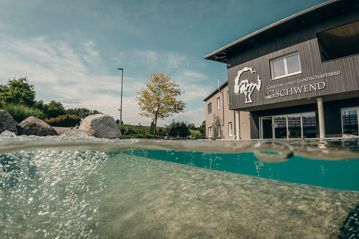 Ein Split-Level-Foto zeigt einen klaren Pool mit Blasen im Vordergrund und ein modernes Gebäude mit einem Baum-Logo und einem Gschwend-Schild im Hintergrund, unter einem teilweise bewölkten Himmel.