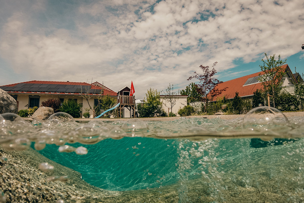 Eine Halbunterwasseransicht eines Pools in einem Hinterhof, mit Blasen auf der Wasseroberfläche und zwei Häusern mit roten Dächern und einem hölzernen Spielgerät im Hintergrund unter einem teilweise bewölkten Himmel.