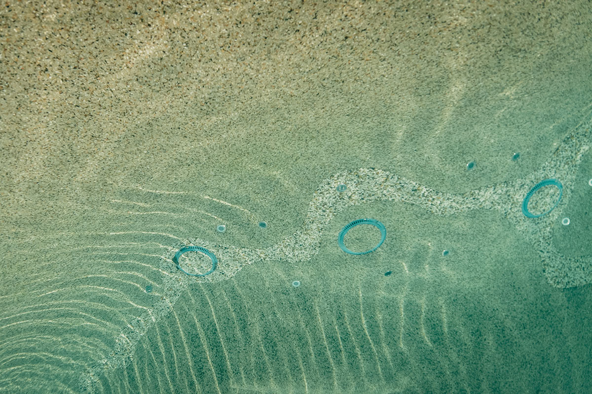 Unterwasseransicht einer sandigen Oberfläche mit sanften Wellen und Sonnenlicht; blaue kreisförmige Poolfliesen sind entlang einer gebogenen Mosaiklinie verstreut.