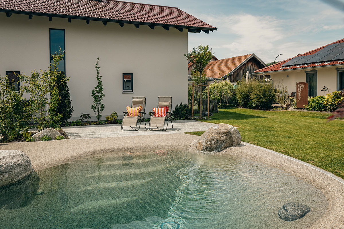 Zwei Liegestühle mit bunten Kissen stehen auf einer Steinterrasse neben einem natürlich wirkenden Pool in einem sonnigen Hinterhof, umgeben von grünem Gras und modernen Häusern mit roten Dächern.