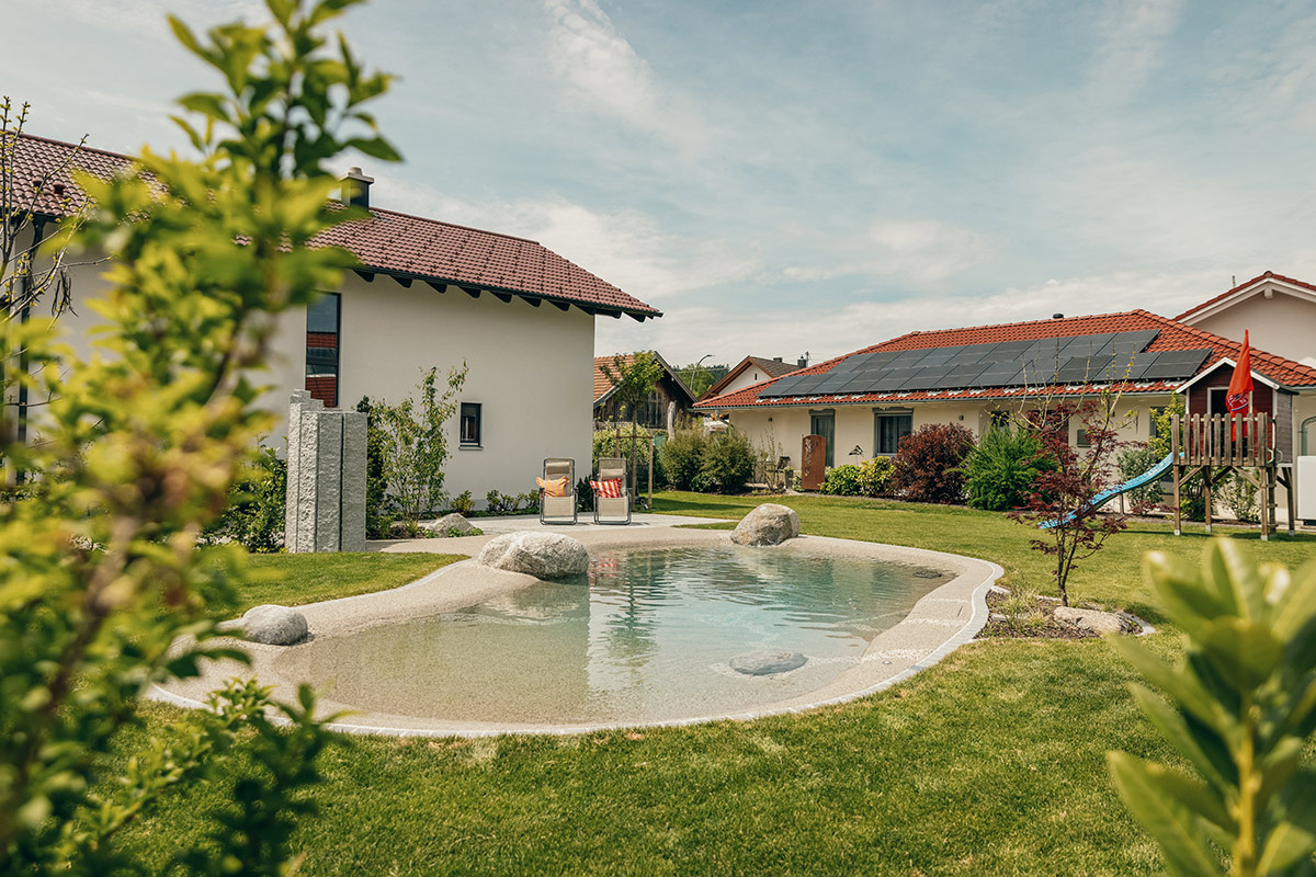 Im Hinterhof befindet sich ein natürlicher Schwimmteich mit Steinen, umgeben von grünem Gras und Pflanzen, in der Nähe von zwei Häusern, eines davon mit Sonnenkollektoren auf dem Dach.