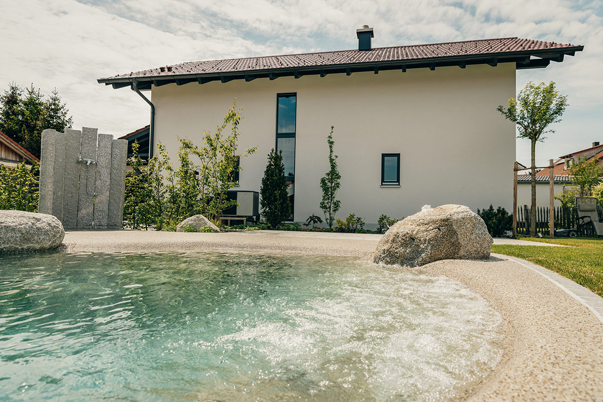 Ein modernes weißes Haus mit rotem Ziegeldach ist hinter einem runden, glitzernden Pool mit Steinen und Rasenflächen zu sehen, unter einem teilweise bewölkten Himmel.