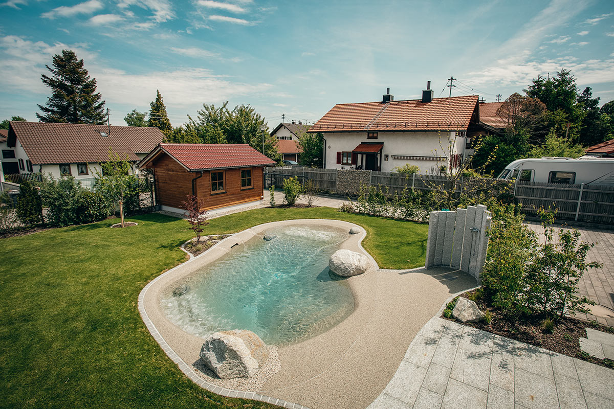 Ein kleiner Garten mit einem natürlichen, lagunenartigen Pool, umgeben von Gras, Steinen und Bäumen, mit Häusern und einem Holzschuppen im Hintergrund. Das Bio.design-Logo ist unten rechts zu sehen.