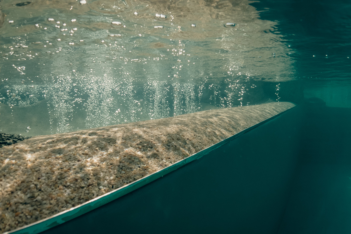 Unterwasseransicht einer Poolwand mit Luftblasen, die an die Oberfläche steigen, und Licht, das vom Wasser reflektiert wird und eine ruhige und klare Szene schafft.