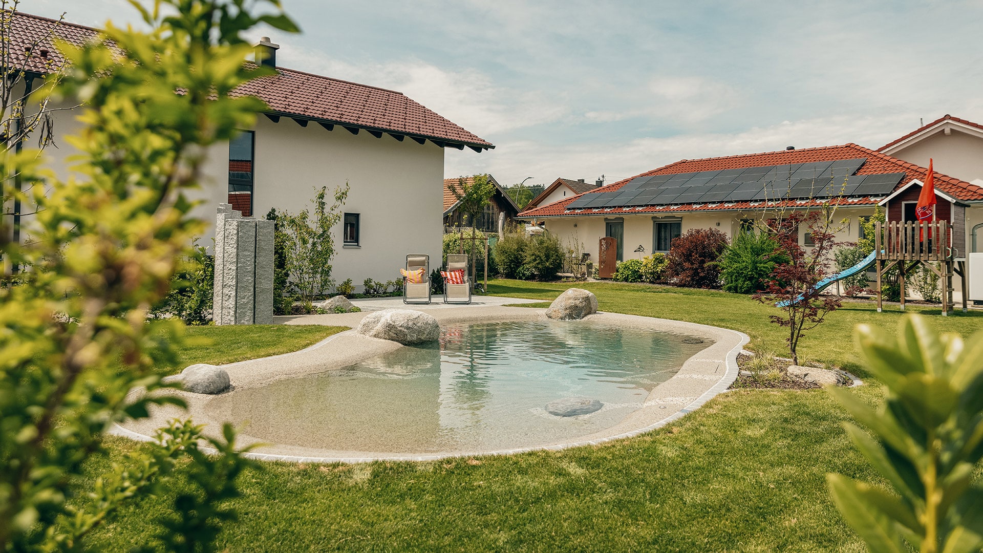 Ein natürlich wirkender Gartenpool mit glatten Rändern und Steinen, umgeben von grünem Gras und Landschaftspflanzen. Zwei Häuser mit roten Dächern, eines davon mit Sonnenkollektoren, stehen im Hintergrund unter einem teilweise bewölkten Himmel.