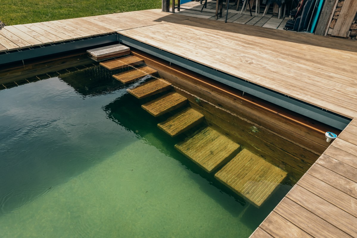 Ein rechteckiges Naturschwimmbecken mit klarem Wasser, einem Holzdeck und einer Holztreppe, die in das Becken führt. Das Deck umgibt den Pool und in der Nähe gibt es grünes Gras.