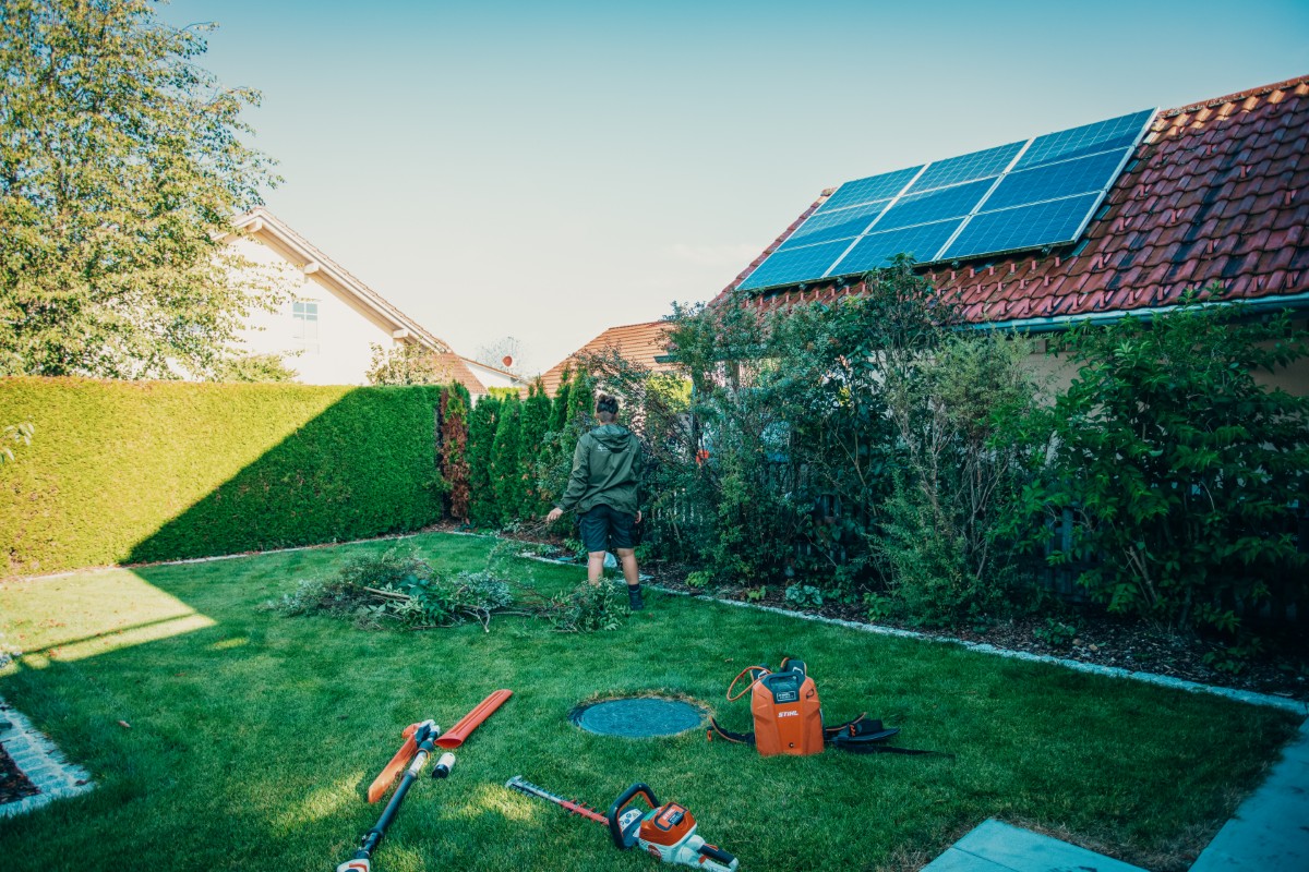 Eine Person steht in einem sonnigen Hinterhof mit gestutzten Büschen und hält Gartengeräte in der Hand. Auf dem Dach des Hauses befinden sich Sonnenkollektoren, und auf dem Rasen liegen verschiedene Gartengeräte.
