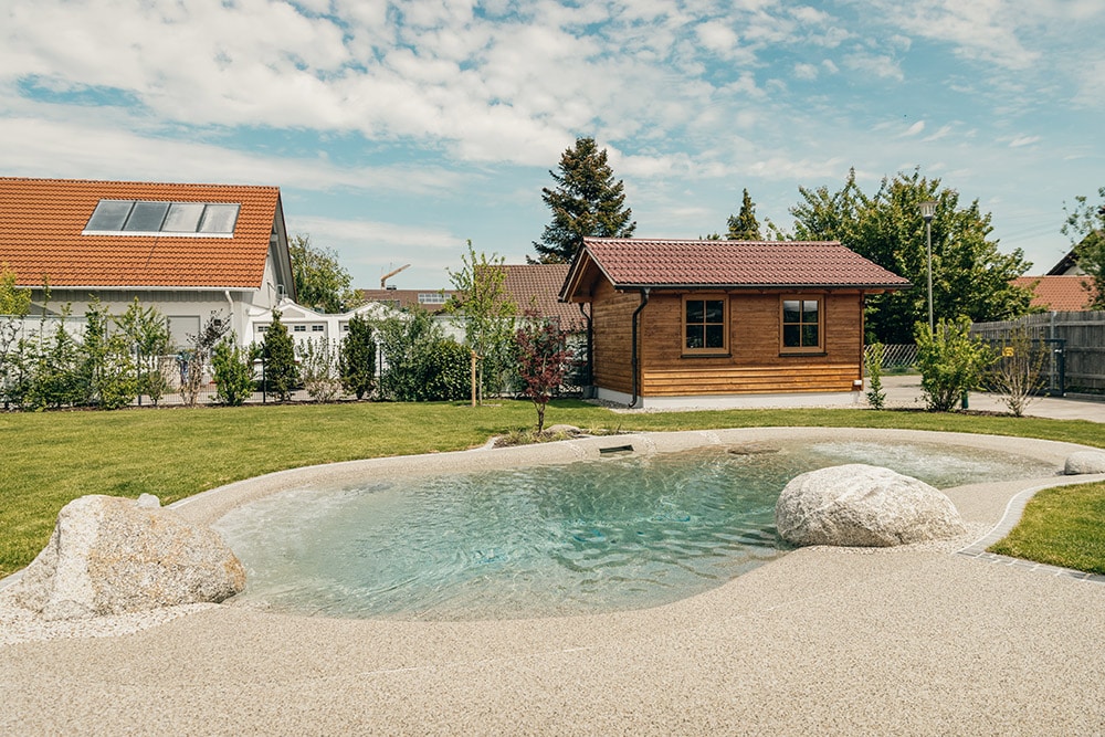 Ein Hinterhof mit einem lagunenartigen Swimmingpool, umgeben von Felsen, grünem Gras, einem kleinen Holzschuppen und einem weißen Haus mit rotem Dach unter einem teilweise bewölkten Himmel.