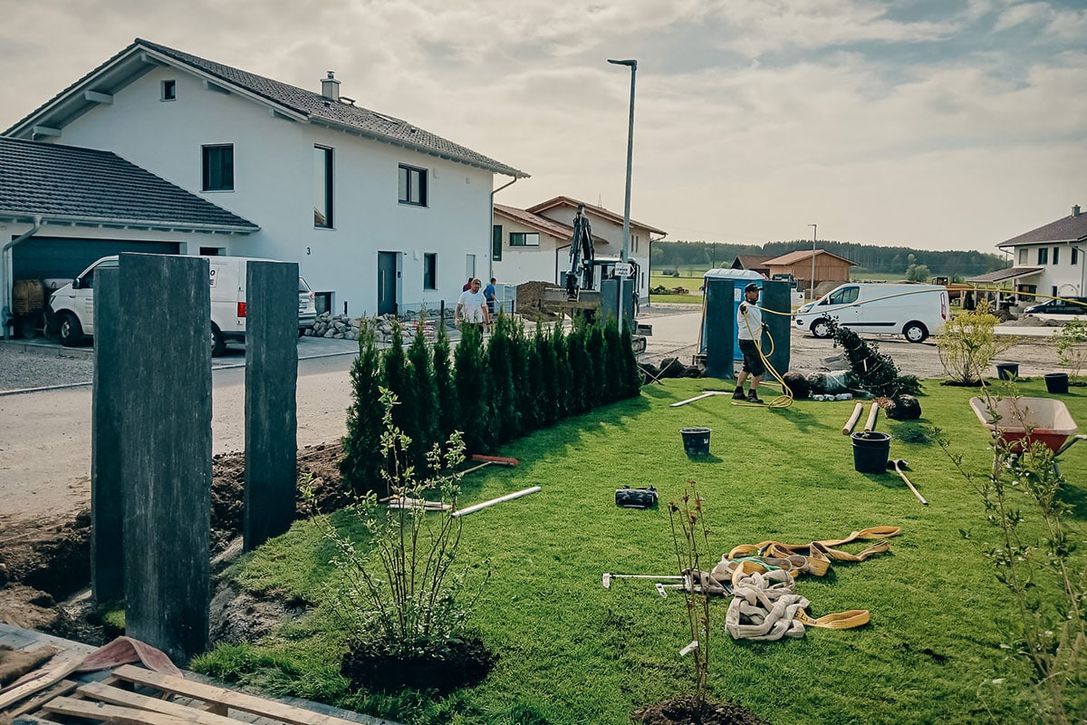 Der Garten- und Landschaftsbau Gschwend gestaltet einen Vorgarten mit jungen Sträuchern und Bäumen auf frischem, grünem Gras. Werkzeuge, Materialien und Versorgungsboxen liegen verstreut herum, während im Hintergrund moderne Häuser und Fahrzeuge zu sehen sind.