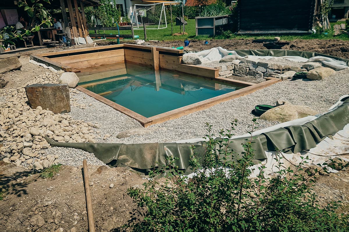 In einem von Garten- und Landschaftsbau Gschwend angelegten Garten befindet sich ein rechteckiges Naturschwimmbecken mit hölzernen Seitenwänden, umgeben von Kies, Steinen und freiliegenden Landschaftsbaumaterialien. Rundherum sind Grünflächen und unvollendete Gartenarbeiten zu sehen.