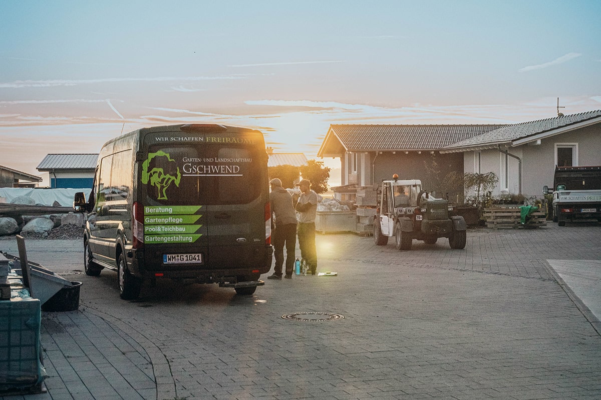 Ein schwarzer Lieferwagen mit der Aufschrift "Garten- und Landschaftsbau Gschwend" steht bei Sonnenuntergang auf einem gepflasterten Parkplatz. Mehrere Personen stehen in der Nähe, im Hintergrund sind ein Traktor und Vorstadthäuser zu sehen, die die Kompetenz des Unternehmens im Landschaftsbau unterstreichen.