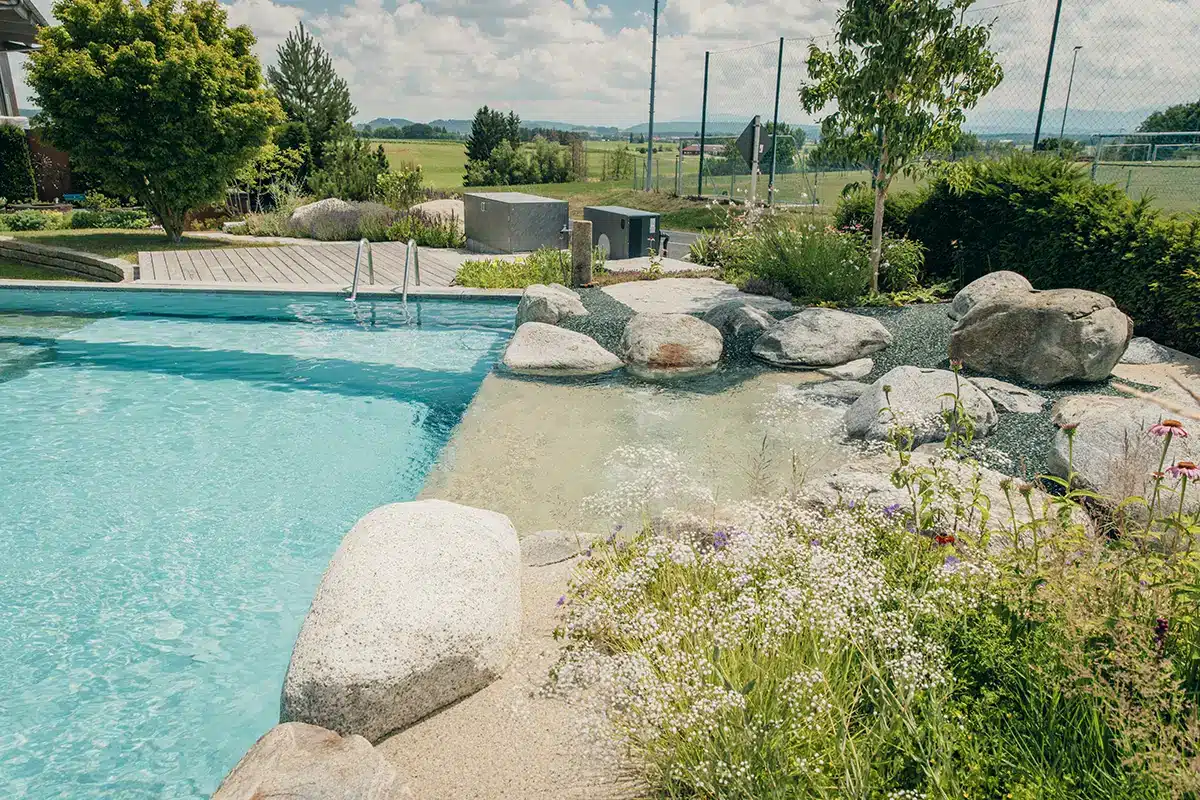 Ein Naturschwimmteich mit klarem, blauem Wasser, Felsen und Wasserpflanzen. Im Hintergrund sind unter einem teilweise bewölkten Himmel eine Holzterrasse, grünes Gras und eine malerische Wiese mit Bäumen und Sportplätzen zu sehen.